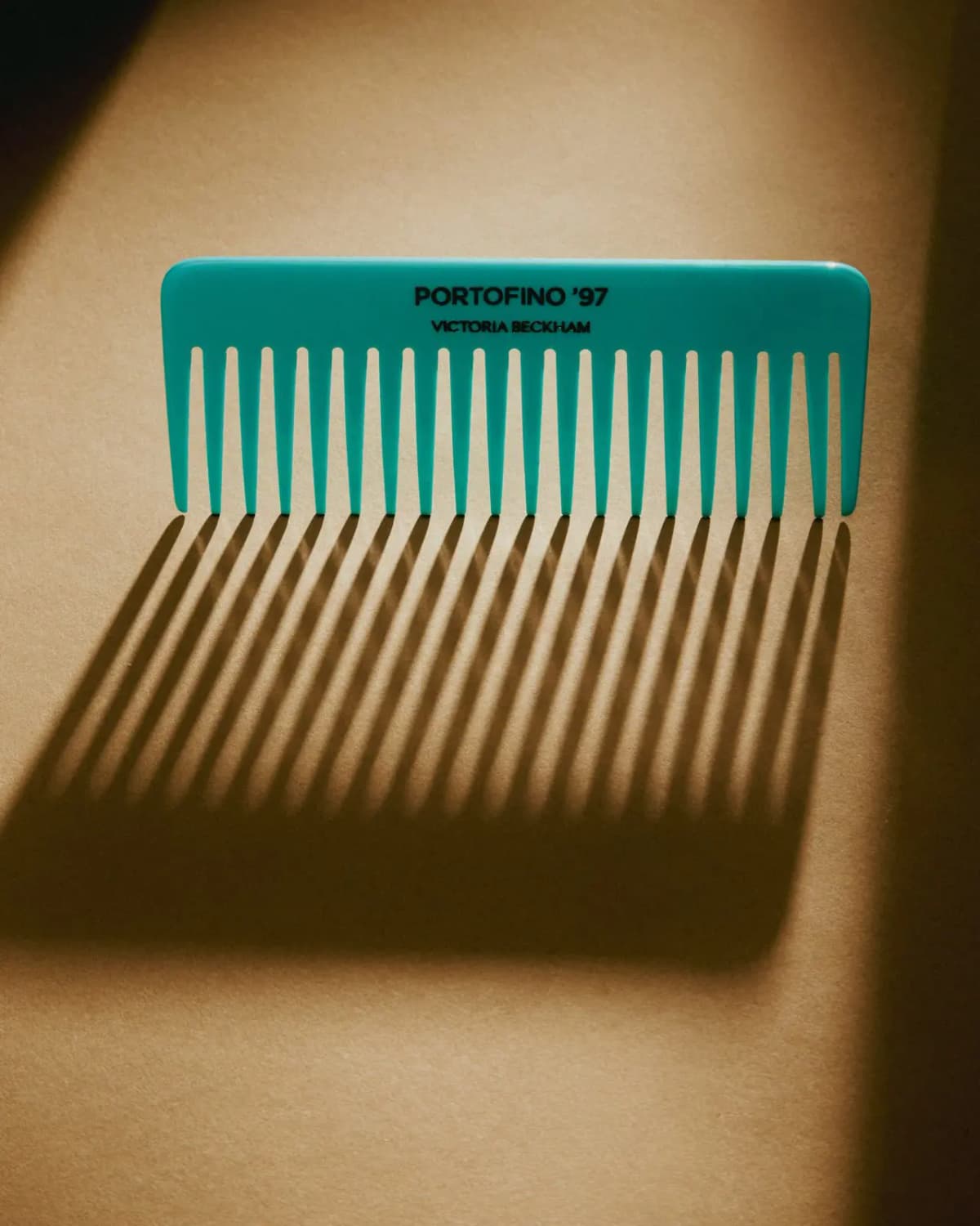 Turquoise wide-tooth Portofino '97 Victoria Beckham comb casting long striped shadows on warm beige paper