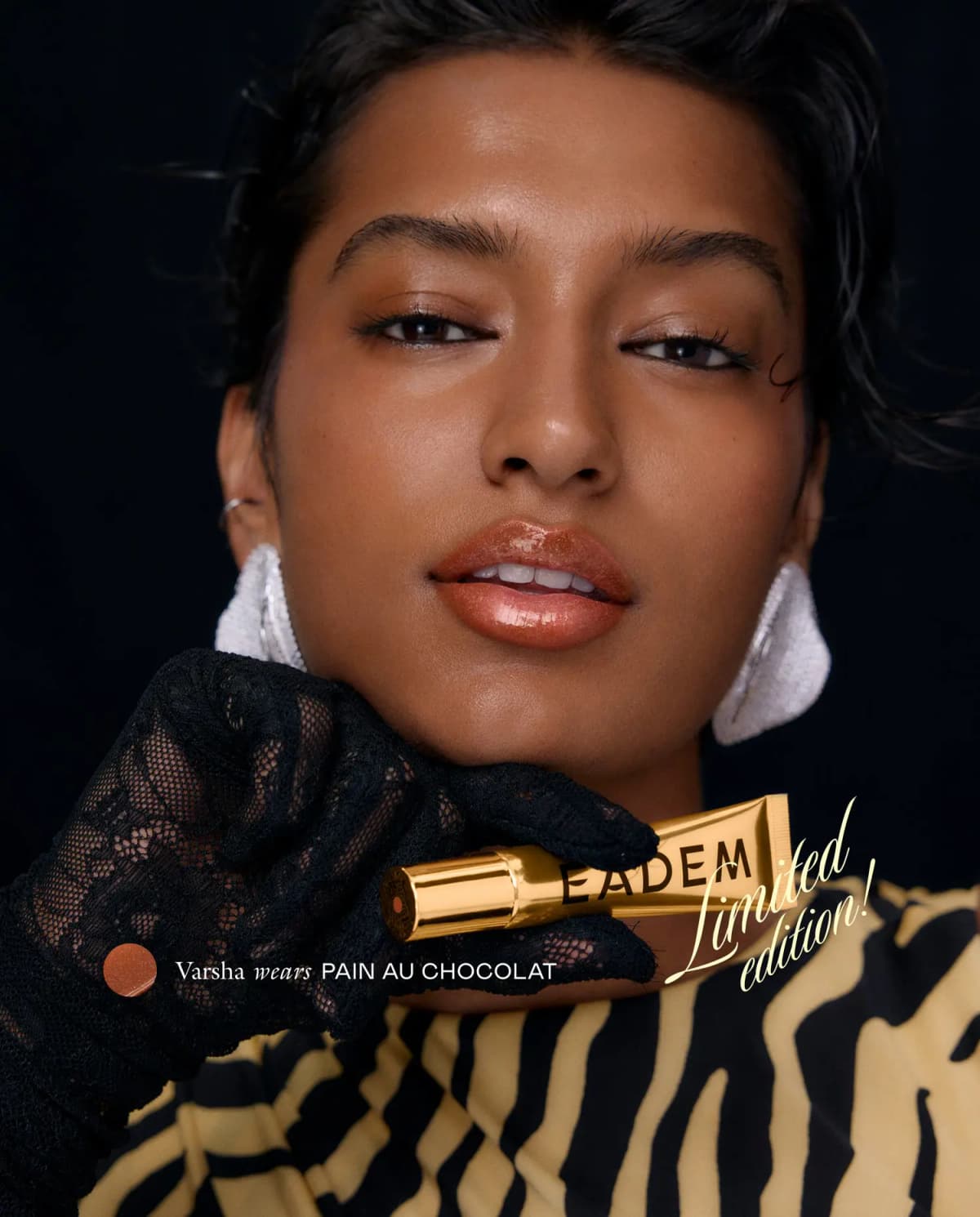 Varsha wears glossy Pain au Chocolat, holding a gold EADEM tube in a black lace glove