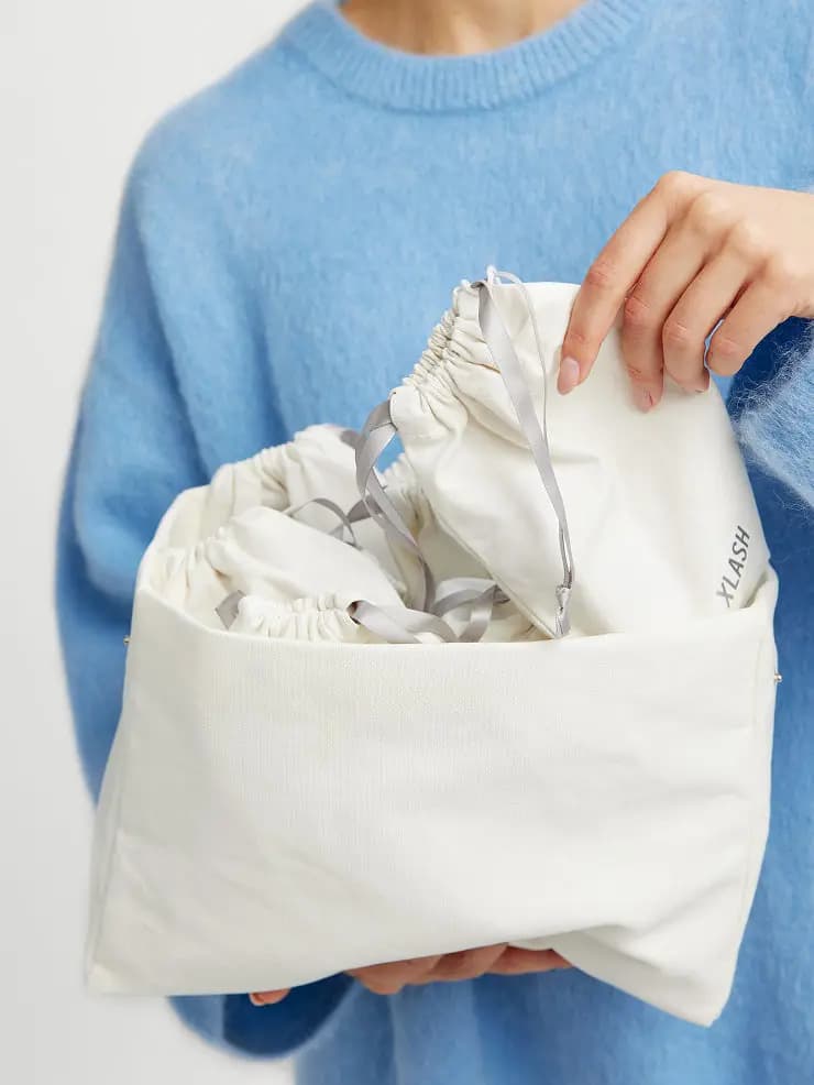 Model in a blue fluffy sweater lifting a cream Xlash drawstring pouch from the cotton beauty bag