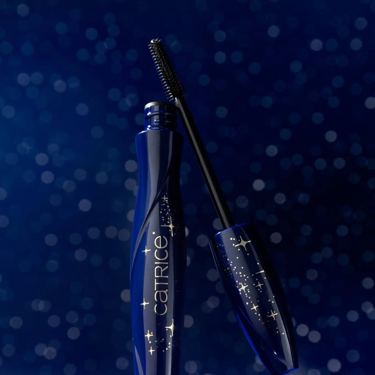 Mascara wand leaning against the open star-printed navy tube on a blue bokeh background