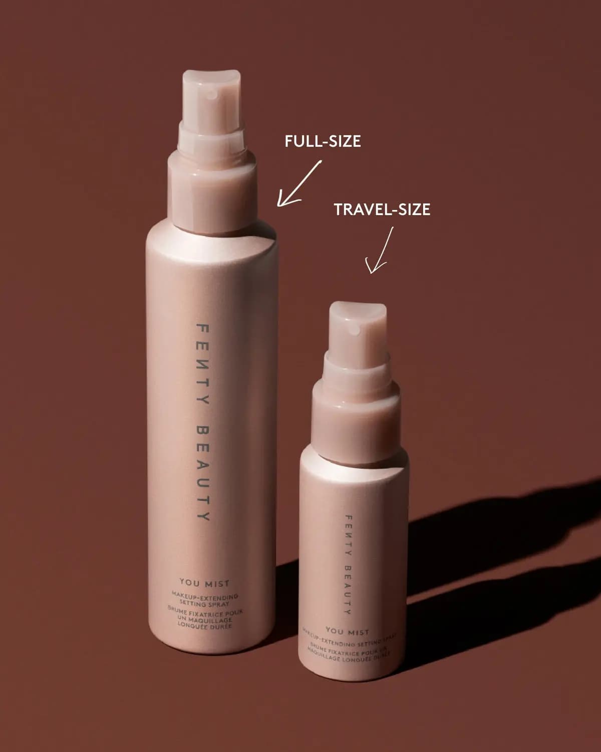 Full-size and travel-size pink Fenty Beauty You Mist setting spray bottles on brown background