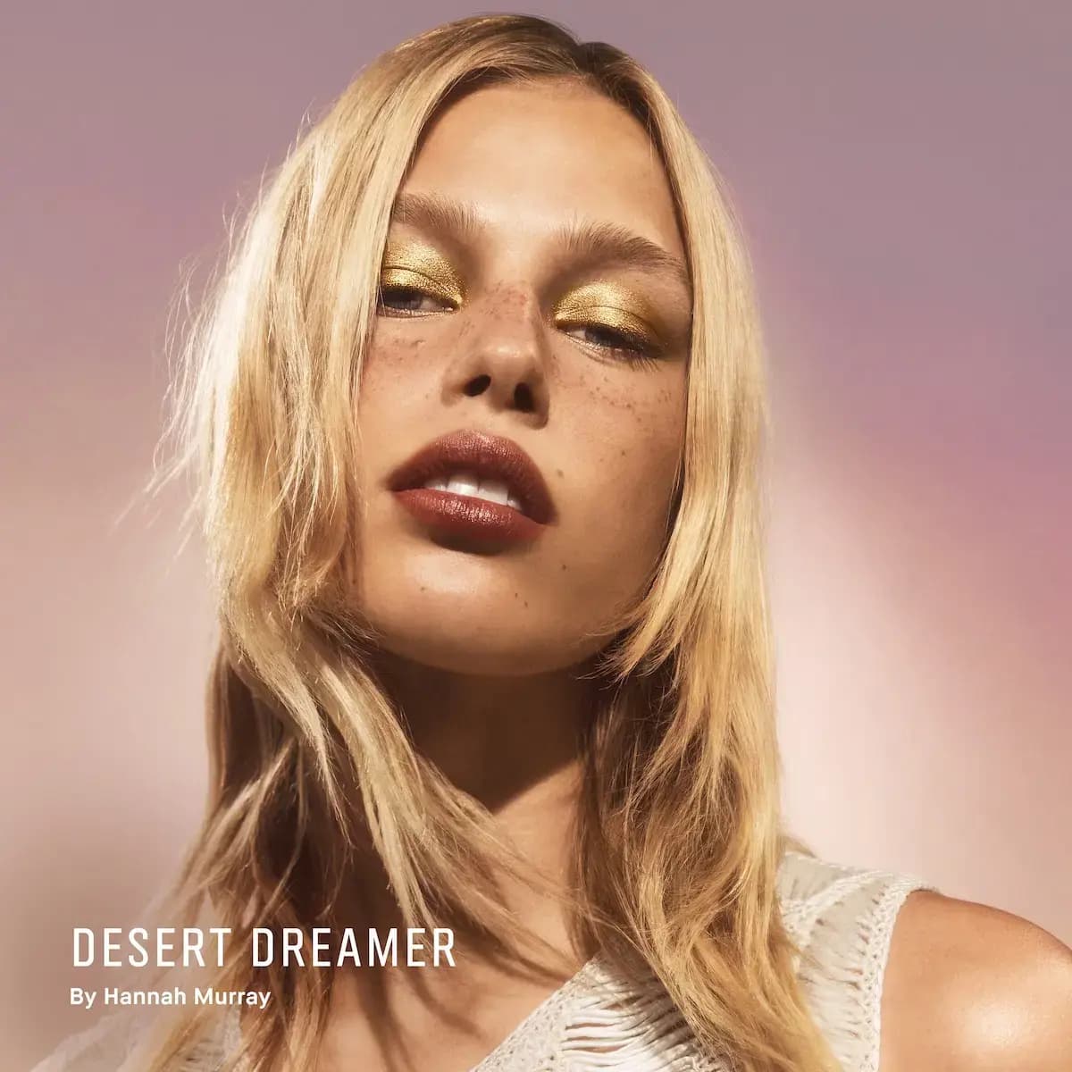 Blonde, freckled model wearing metallic gold eyeshadow and glossy brick-red lips against a pink-mauve backdrop.
