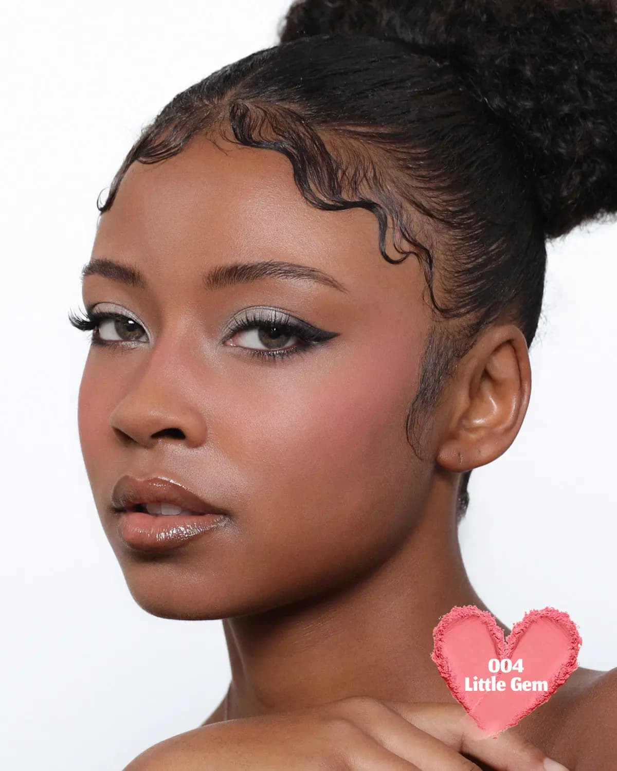 Model with sleek updo wearing a warm coral-pink blush on the cheek, with a heart-shaped 004 Little Gem powder swatch.