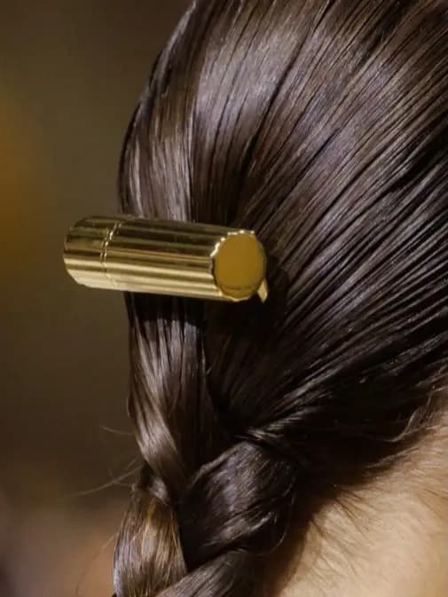 Fluted gold lipstick-shaped hair clip set in sleek dark hair above a braid