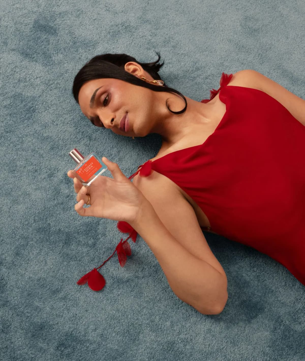 Cyrus Veyssi in a red dress lies on a blue-grey carpet, gazing at a L'Essence Shirin bottle in their hand
