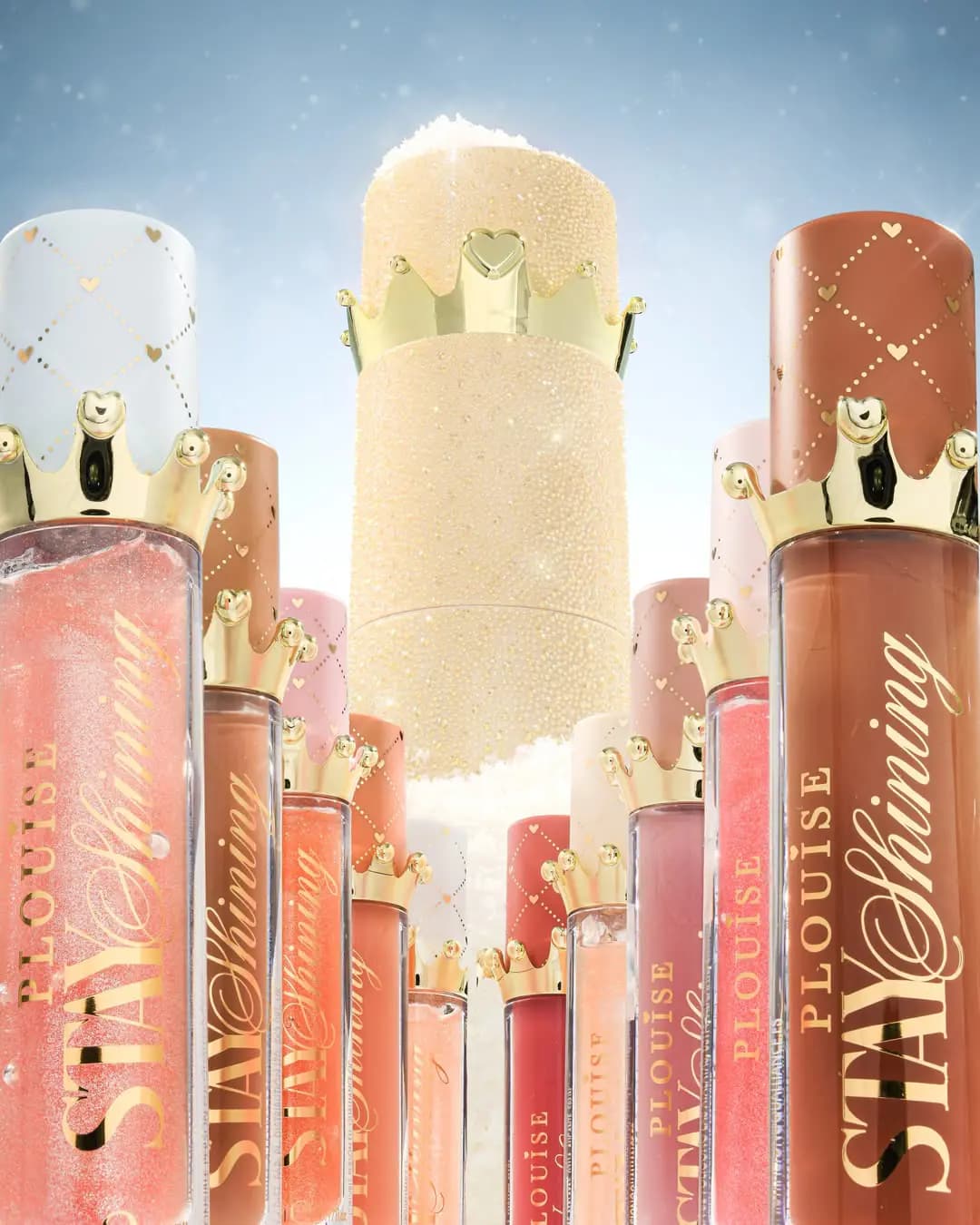 Rows of Stay Shining lip glosses with gold crown caps beside a sparkly gold crown-topped cylinder against a blue sky