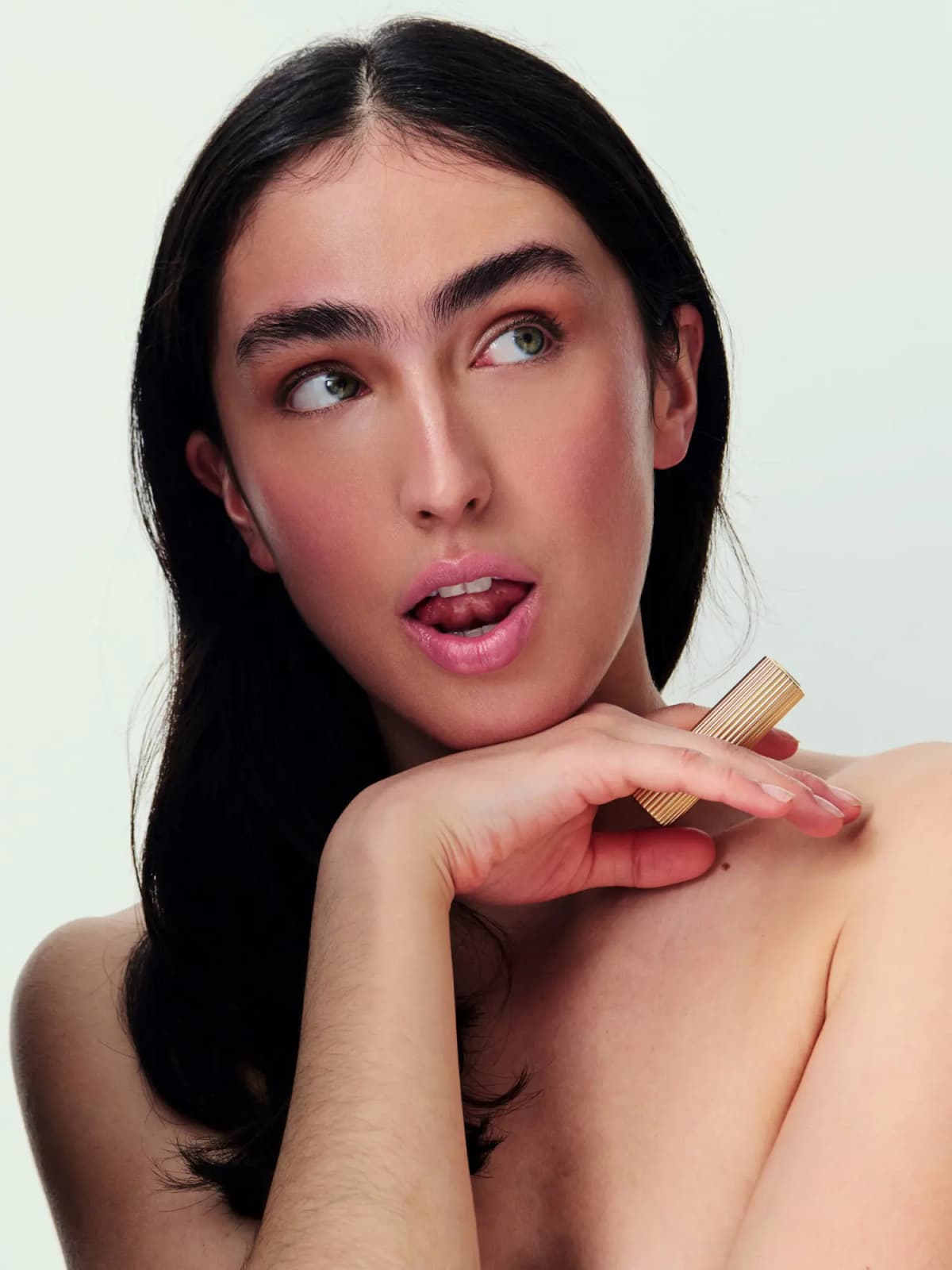 Dark-haired model with soft pink lips and rosy cheeks holds the gold ribbed balm case at her shoulder