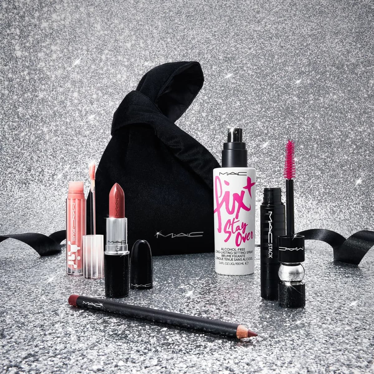 Black knotted pouch with nude lipstick, pink gloss, liner, setting spray and mascara on a silver glitter backdrop