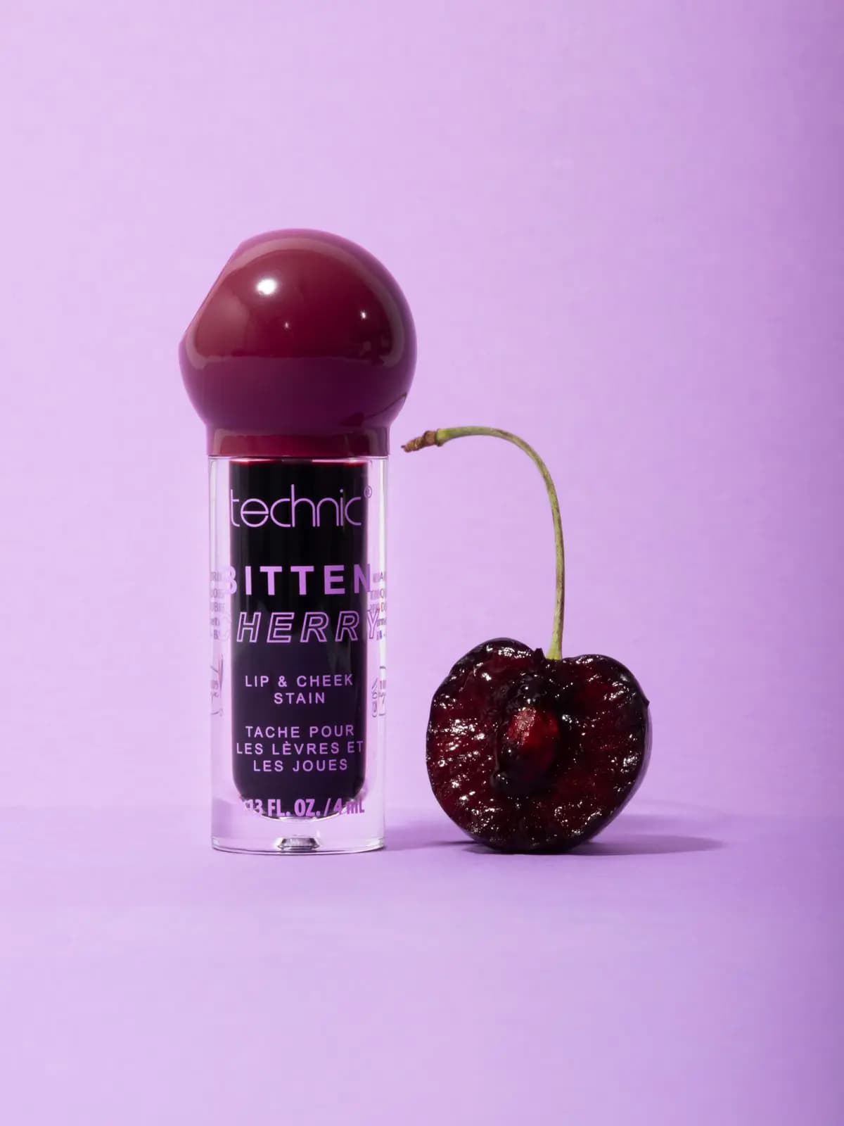 Bitten Cherry Lip & Cheek Stain with a cherry-shaped cap beside a halved juicy cherry on a lilac backdrop