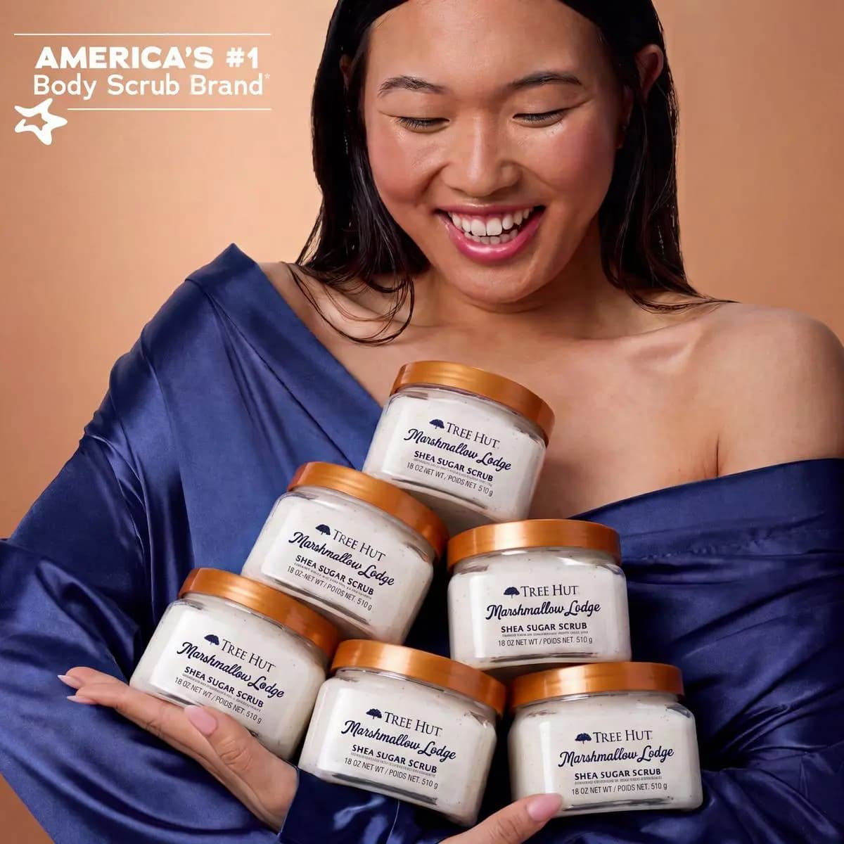 Smiling woman in blue satin top cradling six stacked Marshmallow Lodge Shea Sugar Scrub jars
