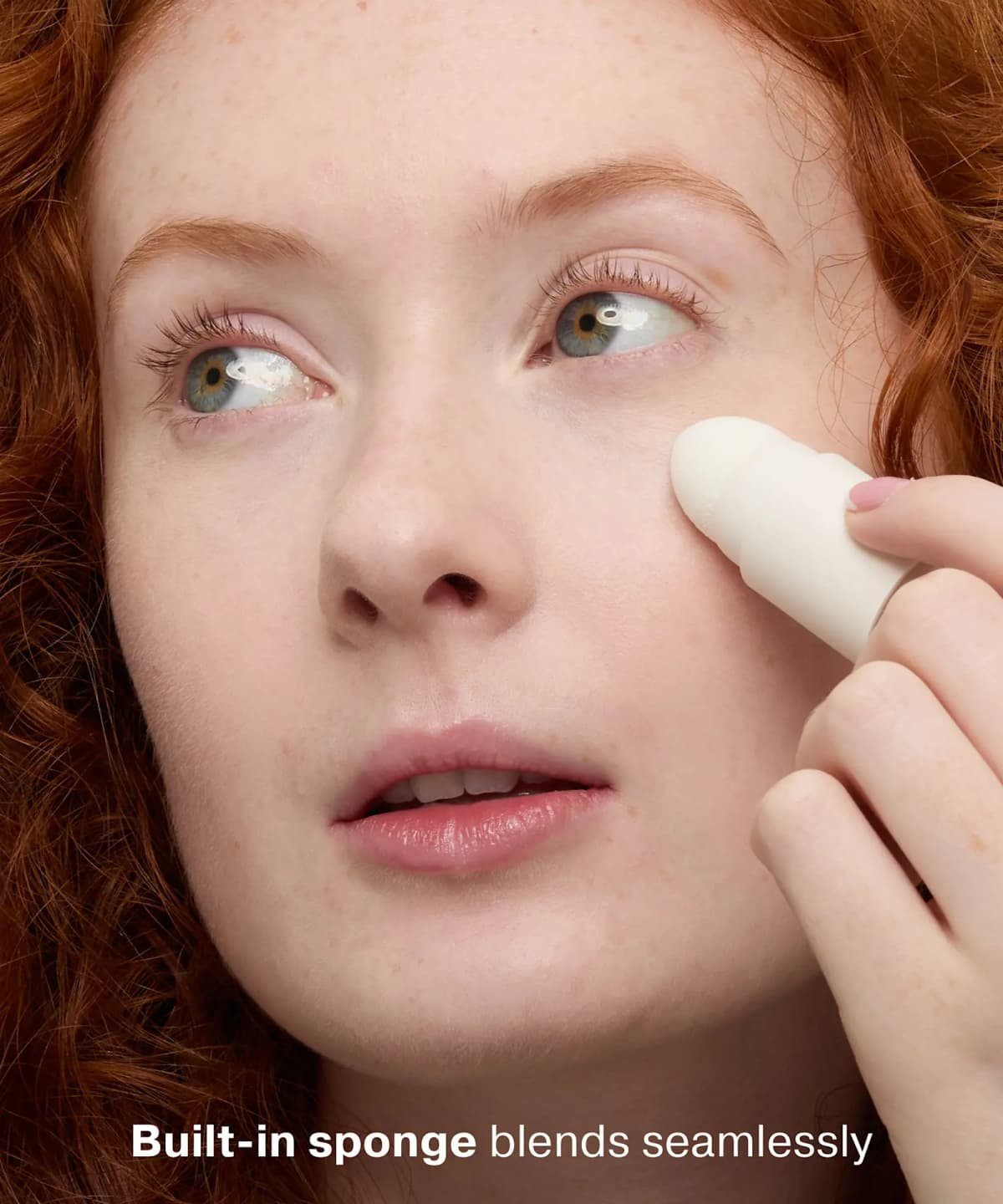 Red-haired freckled model blends concealer under her eye with the built-in white sponge end