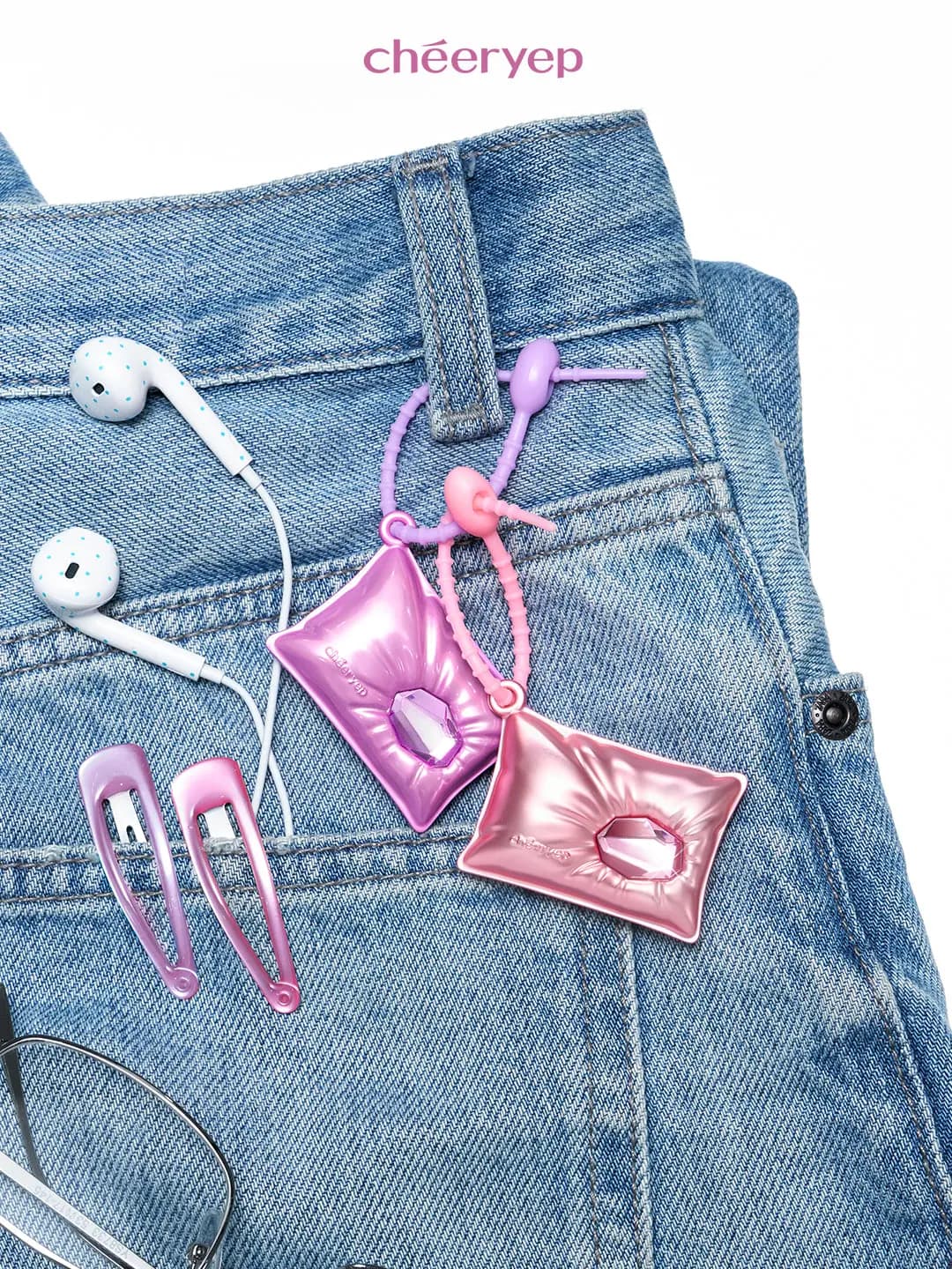 Purple and pink pillow-shaped compacts clipped to a jeans belt loop with earphones, pink hair clips and glasses nearby