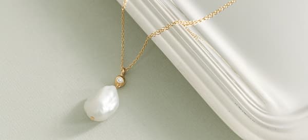 Gold chain necklace with a baroque pearl and small crystal pendant resting on a white ledge