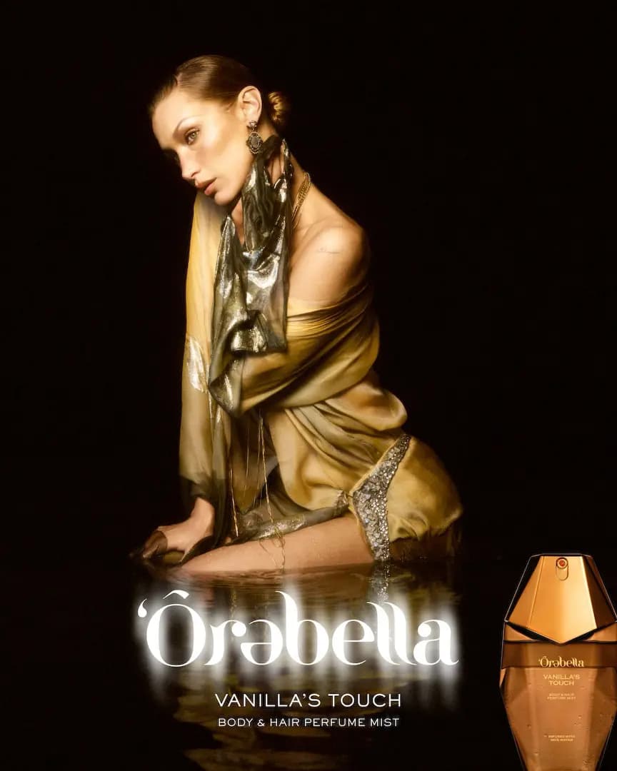 Model in draped gold silk and metallic scarf kneels in dark water beside a bronze Ôrebella Vanilla's Touch mist bottle