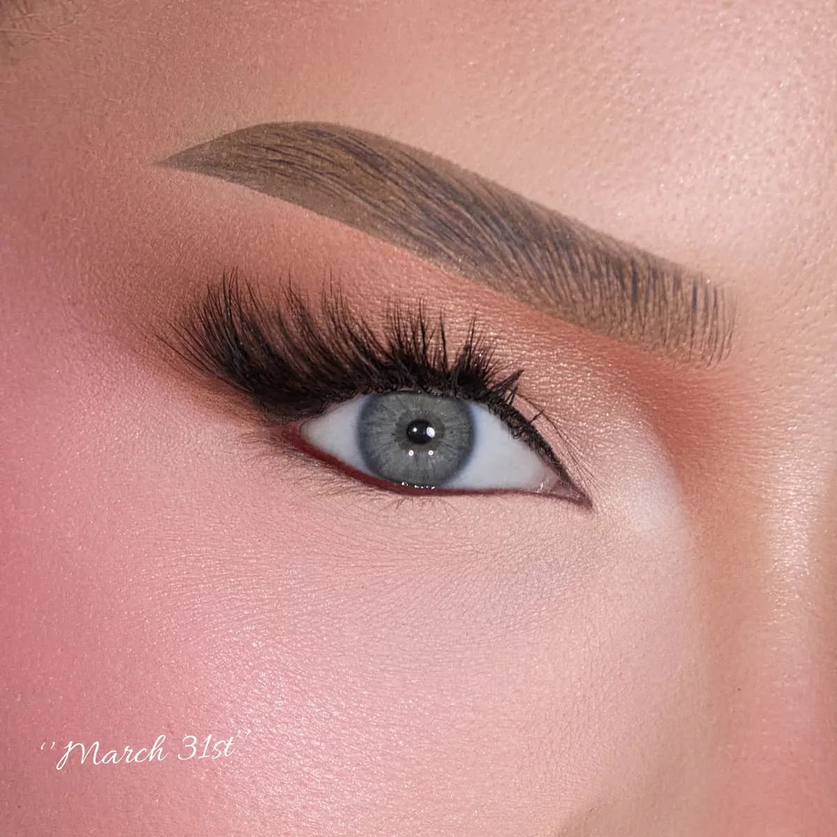 Extreme close-up of a blue eye wearing wispy natural lashes with soft peach shadow, labeled 'March 31st'.
