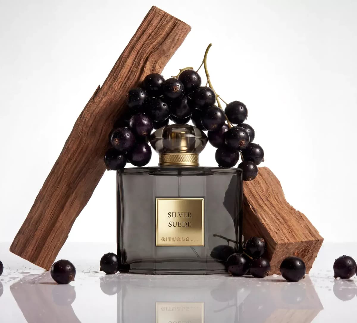 Smoky grey square Rituals Silver Suede bottle with gold label, a blackcurrant cluster and two blocks of wood