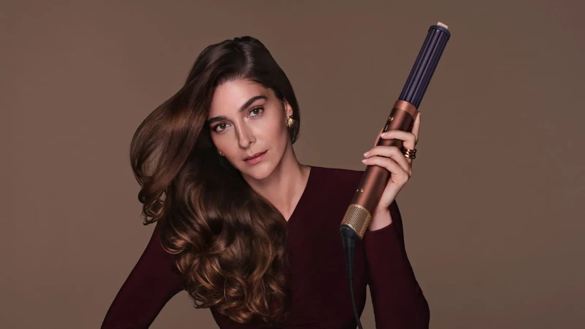 Model with long glossy brunette waves in a burgundy top holding a copper multi-styler with plum barrel