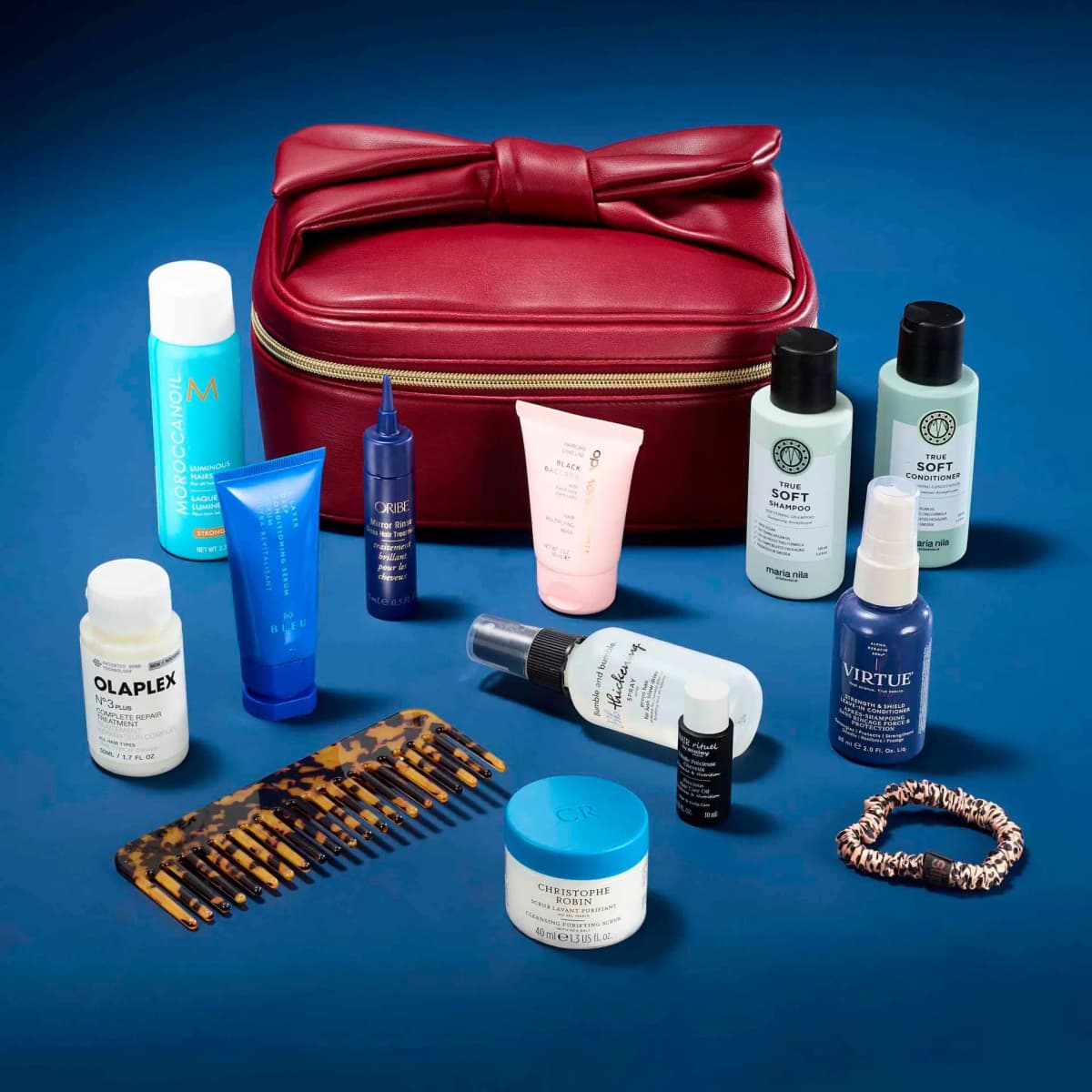 Red bow-top case closed on blue backdrop with Olaplex, Oribe, R+Co Bleu, Christophe Robin minis arranged in front