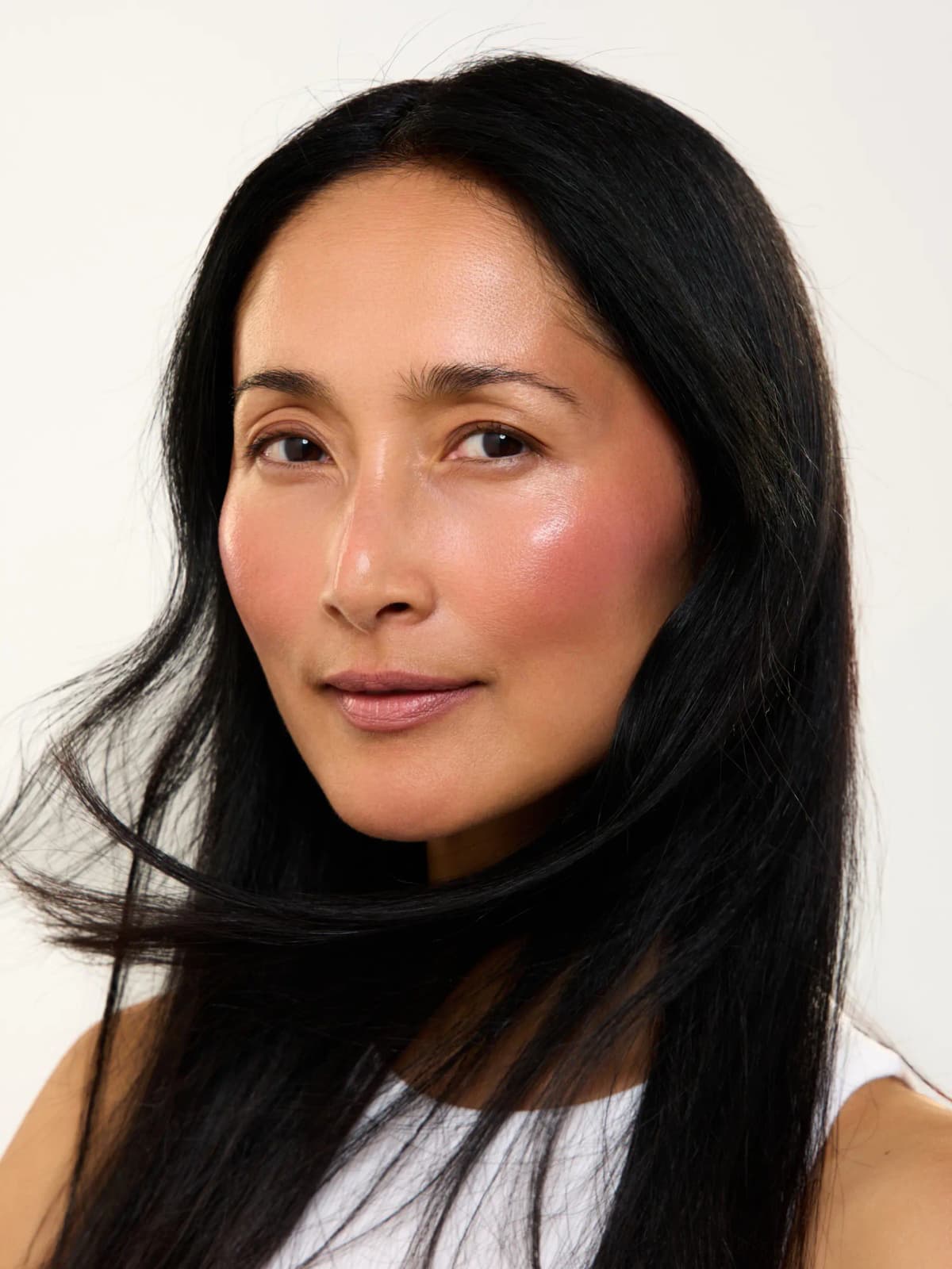 Model with long black hair wearing glossy rosy blush and soft pink lips against a plain white backdrop
