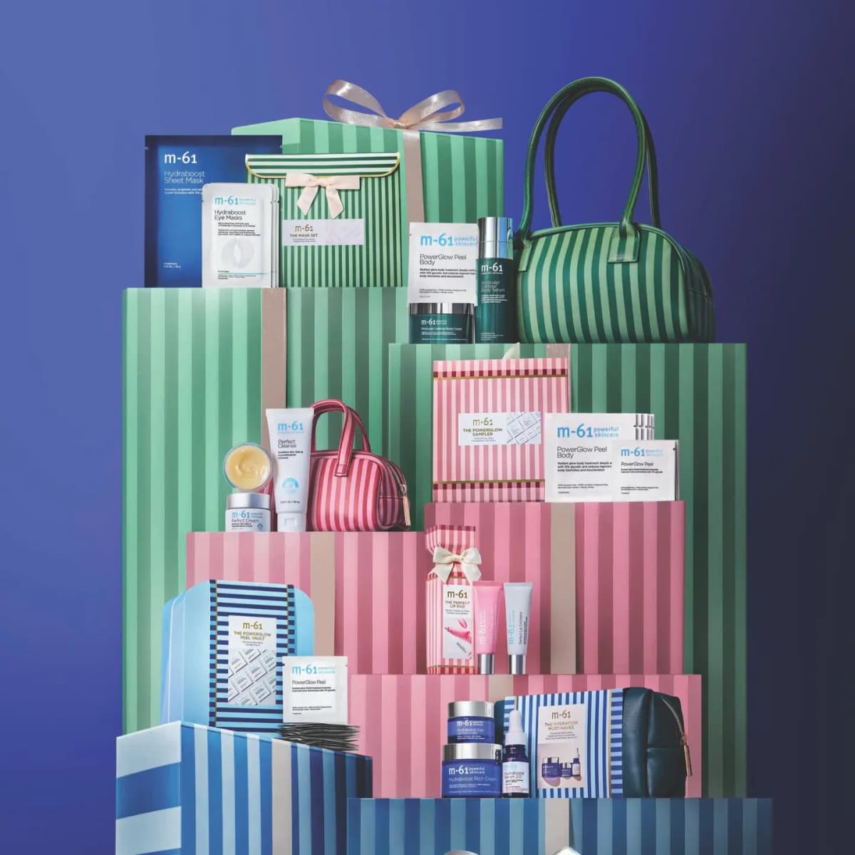 M-61 Holiday 2026 Gift Sets stacked on mint, pink and blue striped gift boxes with striped bags against a deep blue backdrop