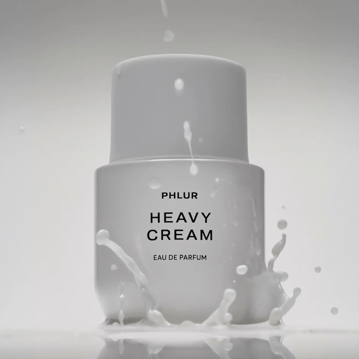White PHLUR Heavy Cream Eau de Parfum bottle with rounded cap amid a splash of milky white liquid on a grey backdrop