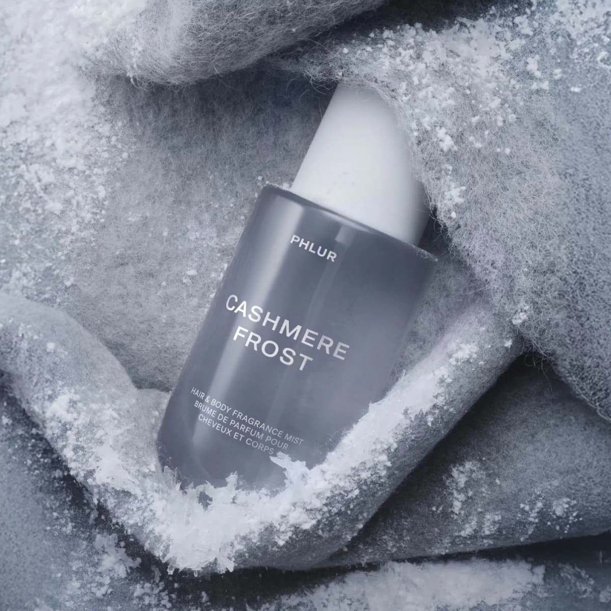 Grey PHLUR Cashmere Frost bottle tilted and nestled in frost-covered grey knit fabric dusted with snow crystals.