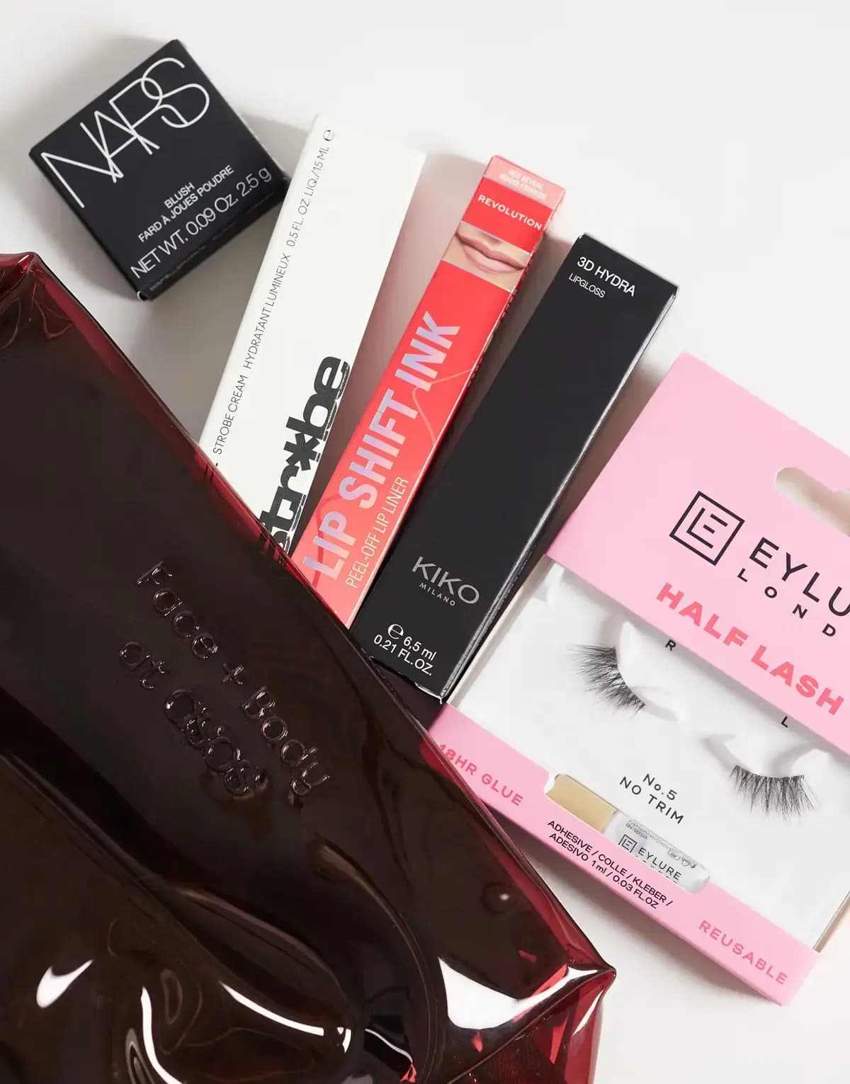 Boxed NARS blush, MAC Strobe Cream, Revolution Lip Shift Ink, KIKO 3D Hydra gloss and Eylure Half Lash beside burgundy bag