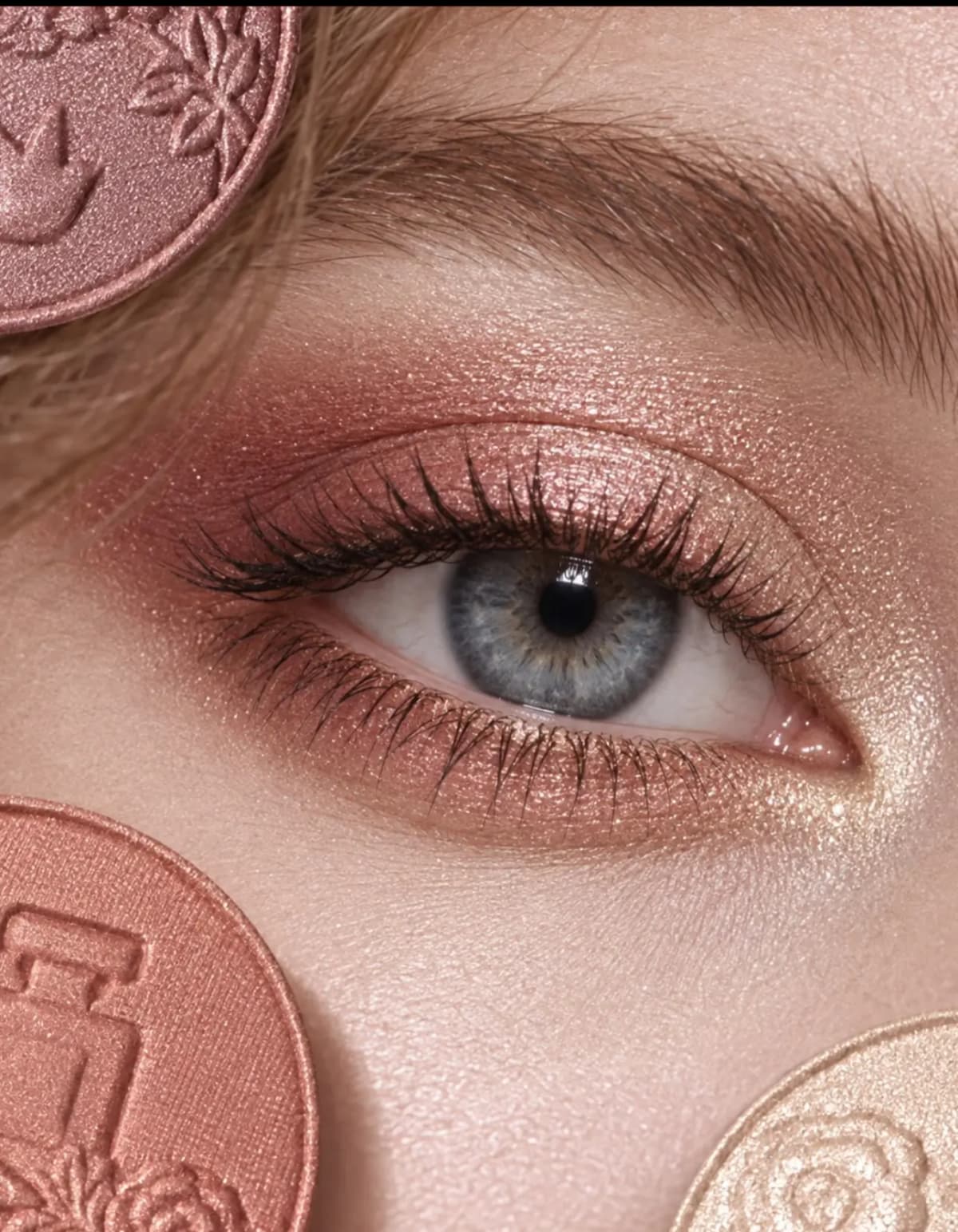 Close-up of a blue eye wearing warm rose gold and champagne shimmer eyeshadow beside coppery pink pressed pans.