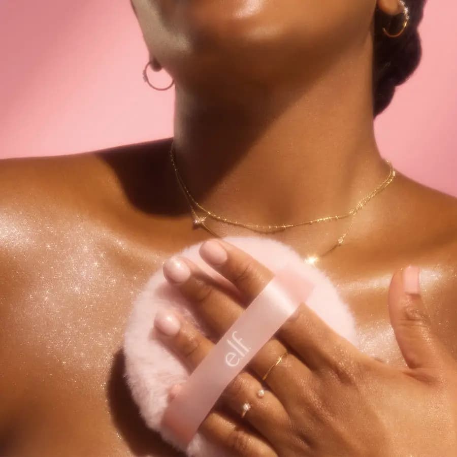Hand pressing the pink puff onto deep-toned chest and shoulder, leaving sparkly glitter, with gold jewellery