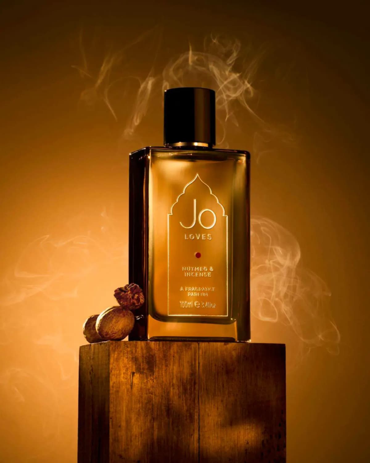 Jo Loves Nutmeg & Incense 100ml bottle on a wooden block with whole nutmegs and swirling smoke on a golden backdrop