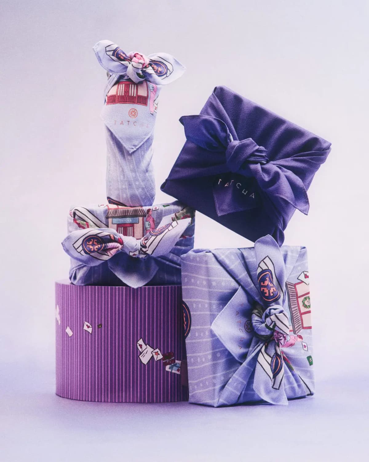 Stack of Tatcha gifts wrapped in illustrated lilac furoshiki and a deep purple cloth, atop a striped purple round box