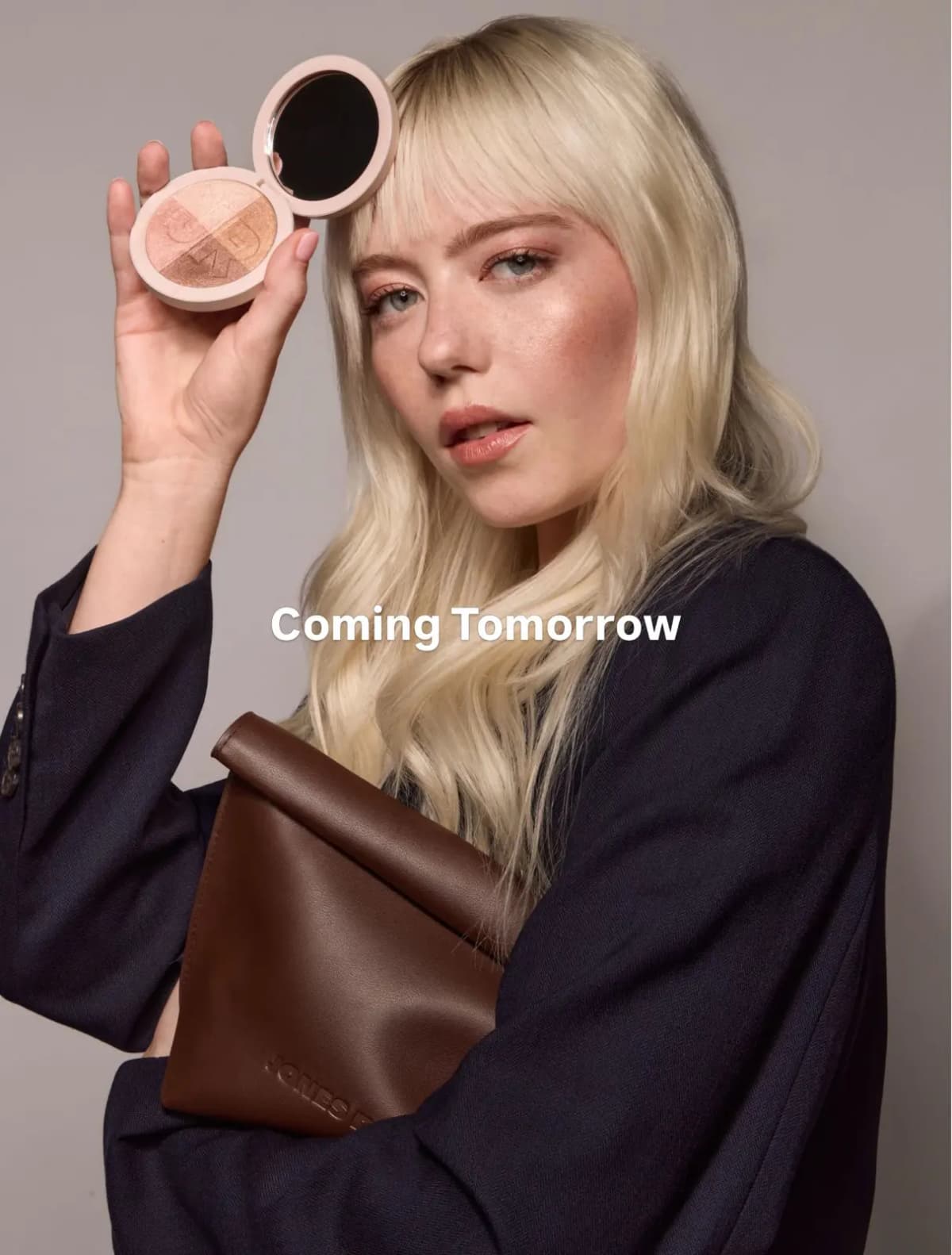 Blonde model with bangs holds open Silky Highlighting Powder compact and brown rolltop clutch, captioned Coming Tomorrow