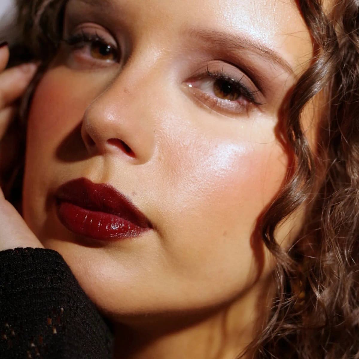 Close-up of a model with curly hair and hazel eyes wearing a glossy dark wine lip, hand resting against her cheek