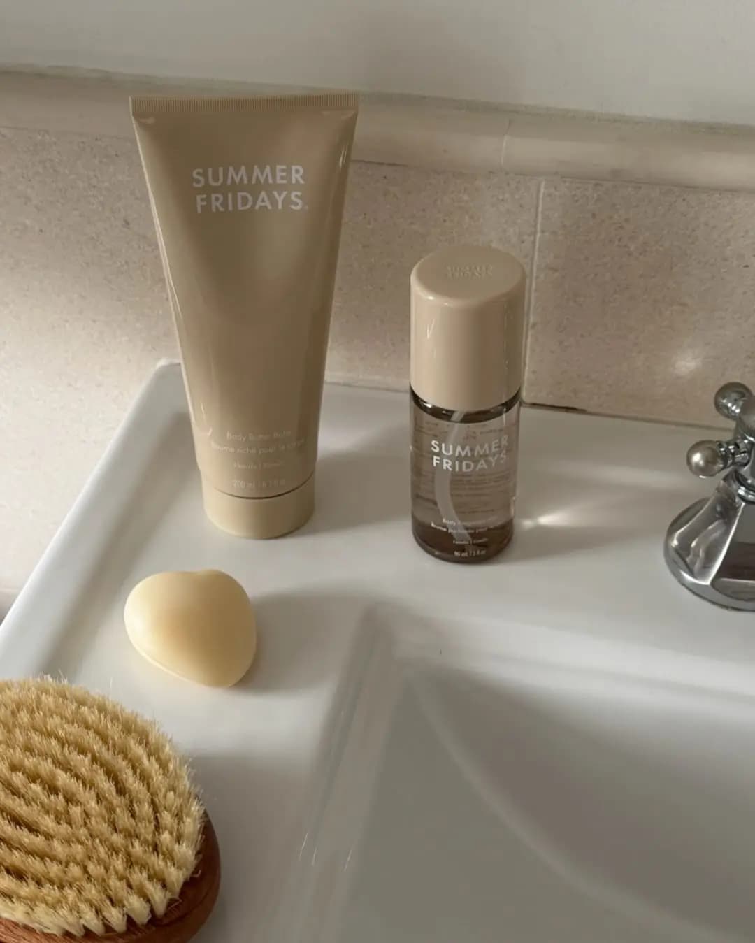 Beige Vanilla Body Butter Balm and Body Fragrance Mist on a white sink with a heart-shaped soap and a bristle brush.