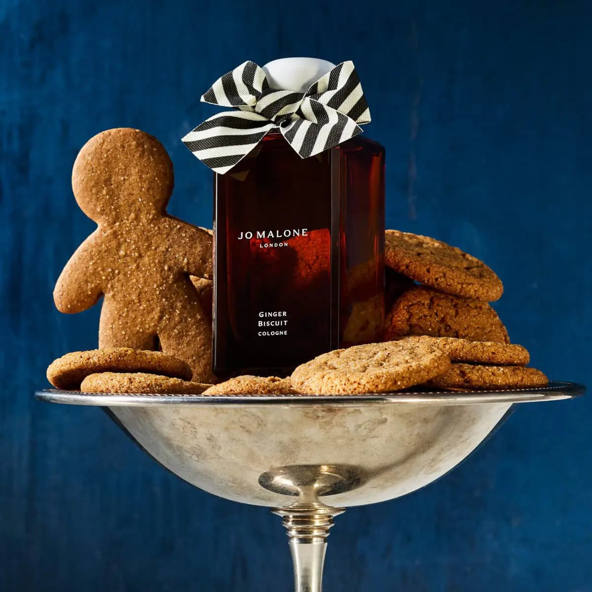 Bottle nestled in a silver pedestal bowl filled with ginger cookies and a gingerbread man against a deep blue backdrop.