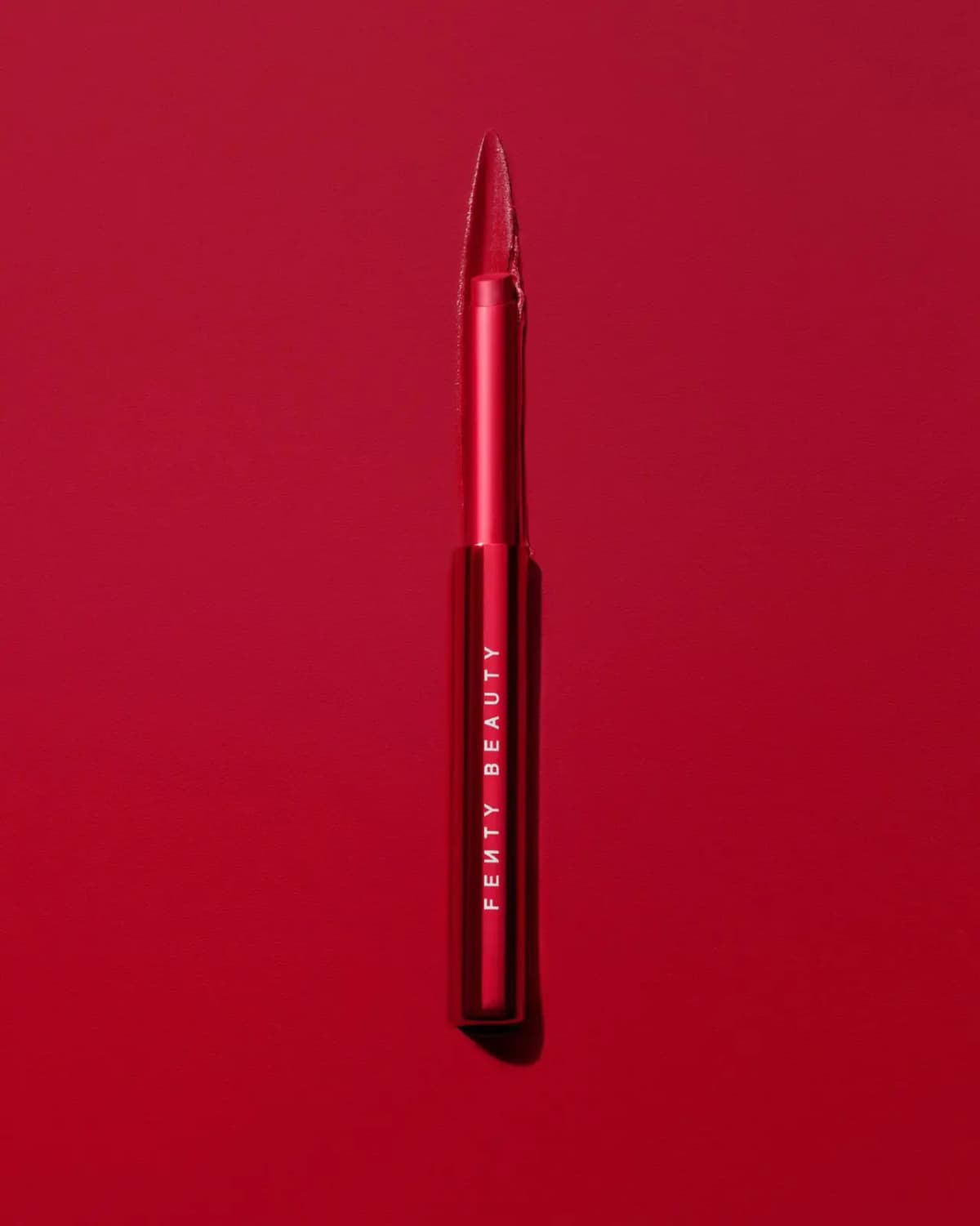 Fenty Beauty Stunna Stix Soft-Glide Matte Lipstick in a red metallic tube on a red backdrop with a matching lipstick swipe