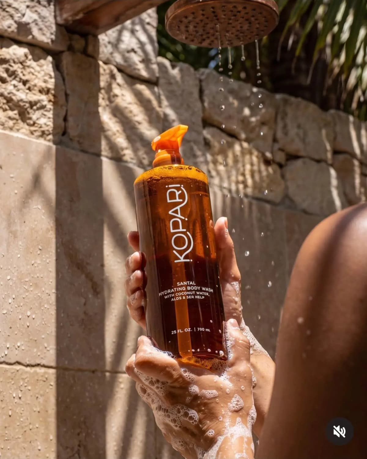 Hands lathered in suds hold the amber Kopari body wash bottle beneath a running outdoor shower head against stone.