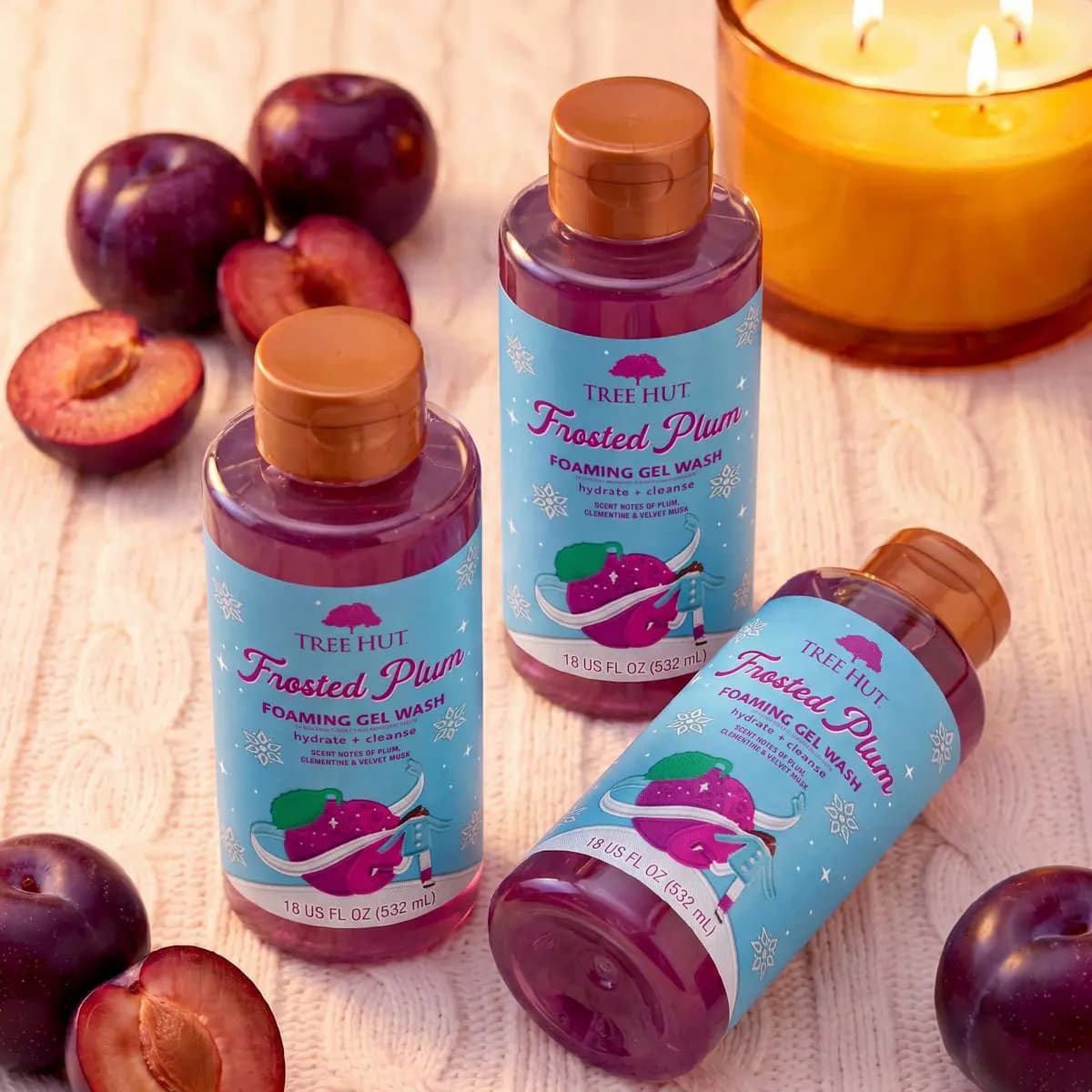 Three Frosted Plum Foaming Gel Wash bottles in translucent purple with fresh plums and a lit amber candle