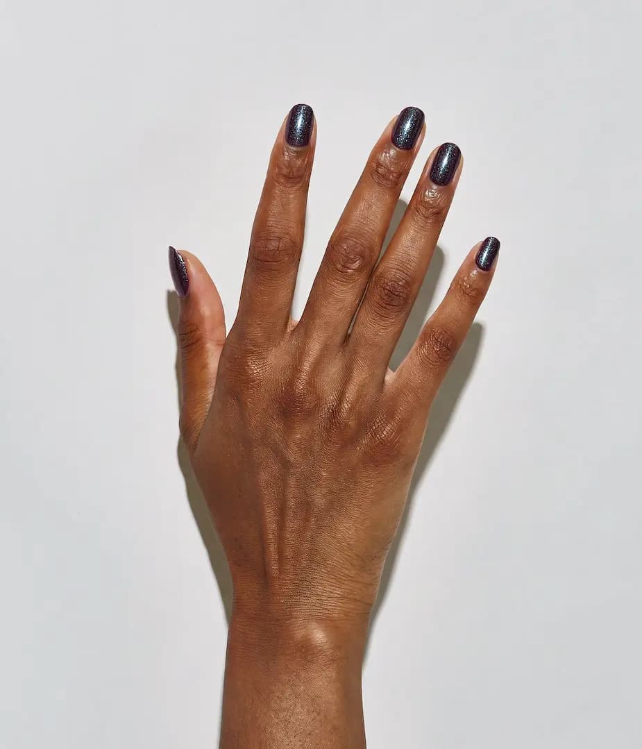 Deep-skinned hand with short nails in a dark polish flecked with blue-green shimmer against a white wall