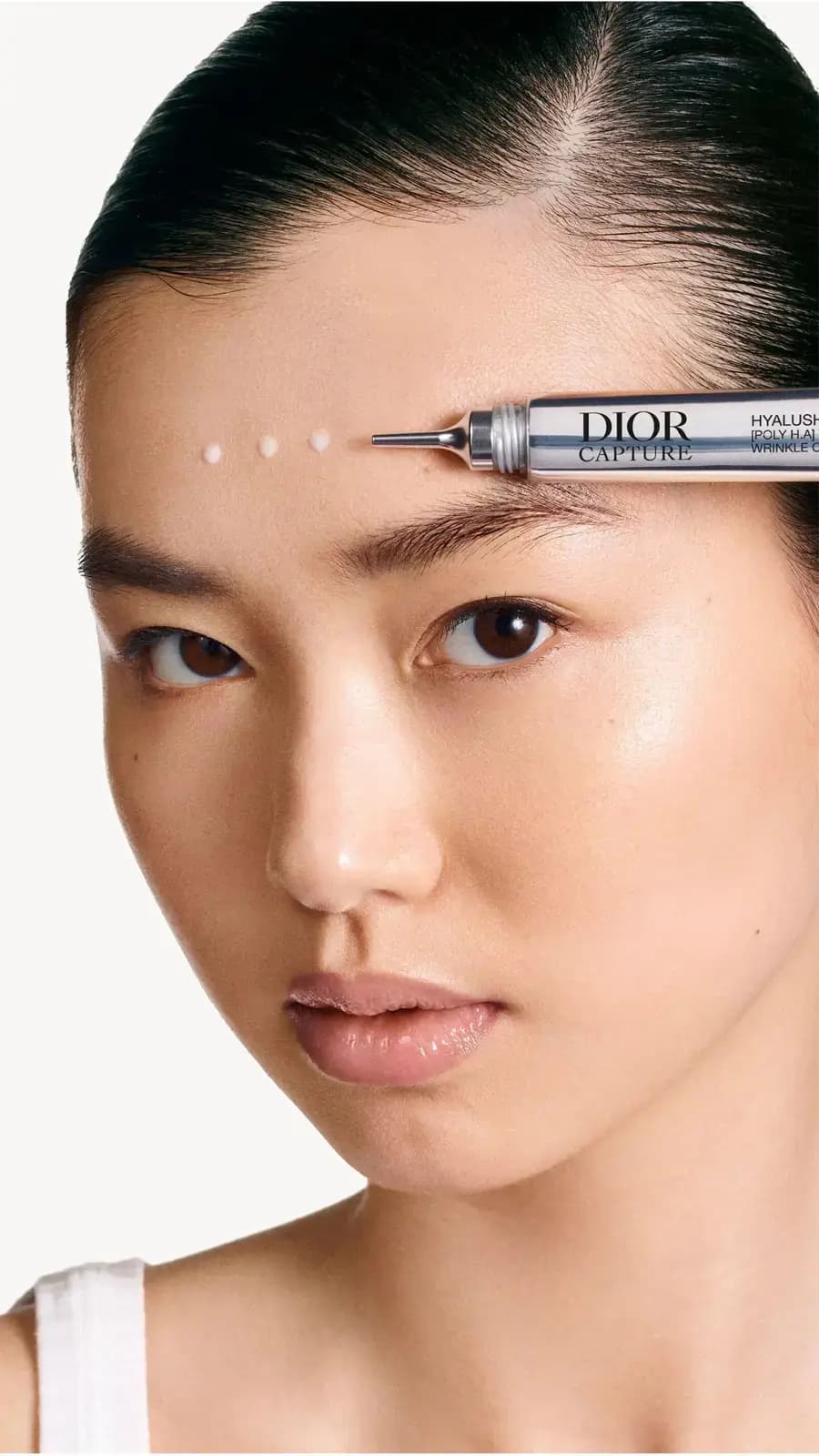 Model with dark slicked-back hair as the applicator places three white dots of product above her eyebrow