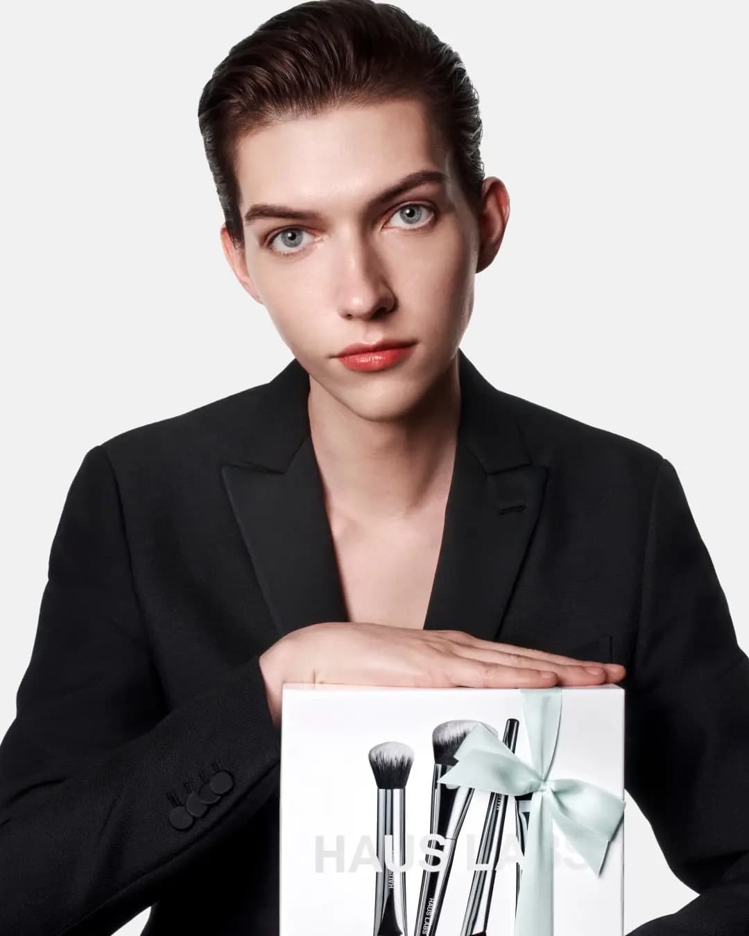 Model in a black blazer resting a hand on a Complexion Brush Set box tied with a mint ribbon