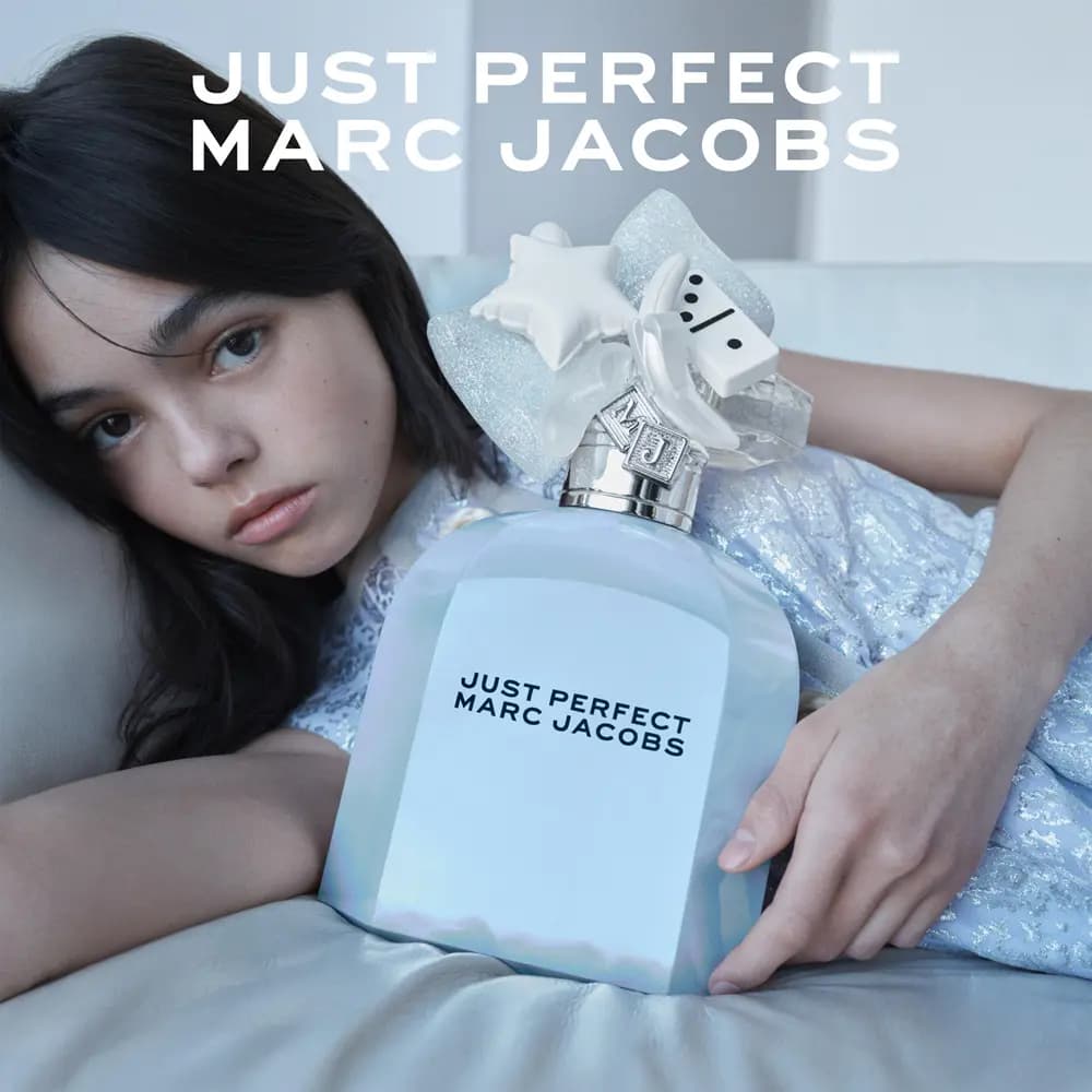 Dark-haired model lying on a sofa hugging a large Just Perfect Marc Jacobs bottle under the brand title
