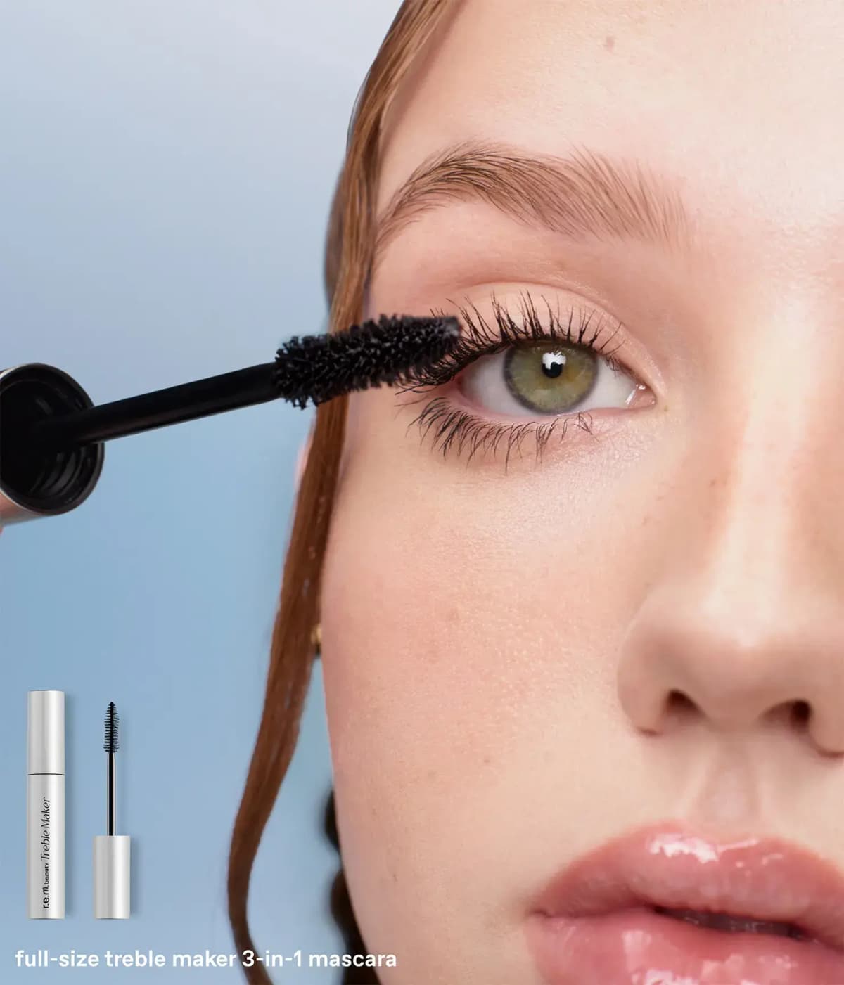 Close-up of mascara wand coating upper lashes on a green-eyed model with red hair, silver tube inset