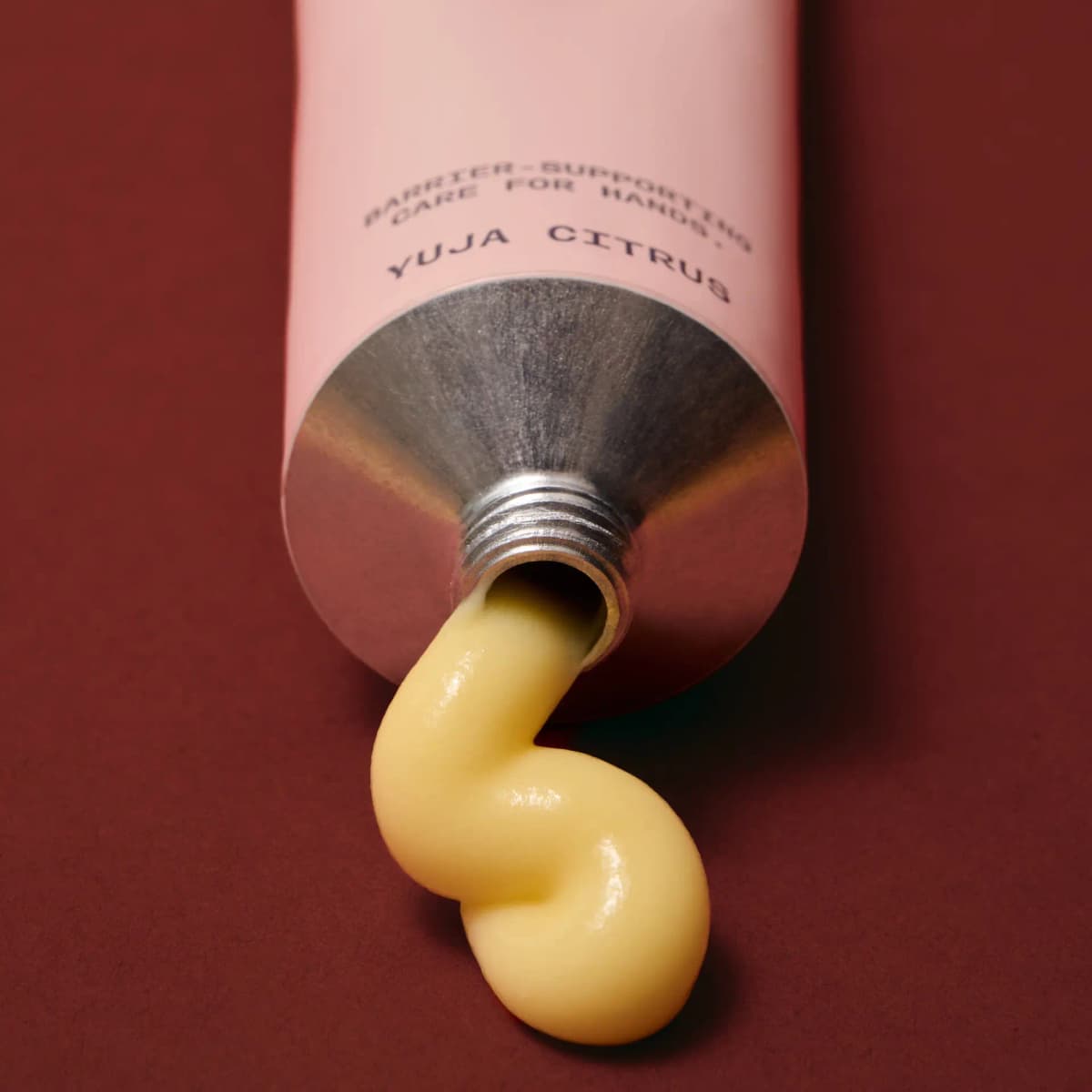 Close-up of pale yellow cream squeezed from the open aluminium neck of a pink Yuja Citrus tube on a red surface