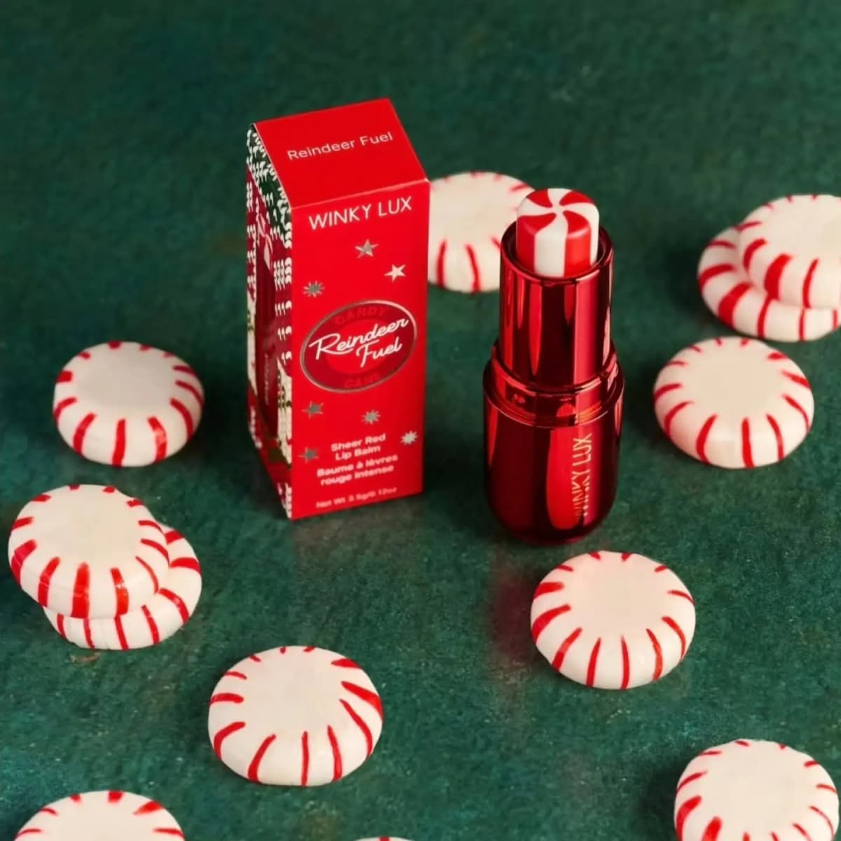 Reindeer Fuel lip balm beside its red Winky Lux box, surrounded by peppermint candies on a green surface
