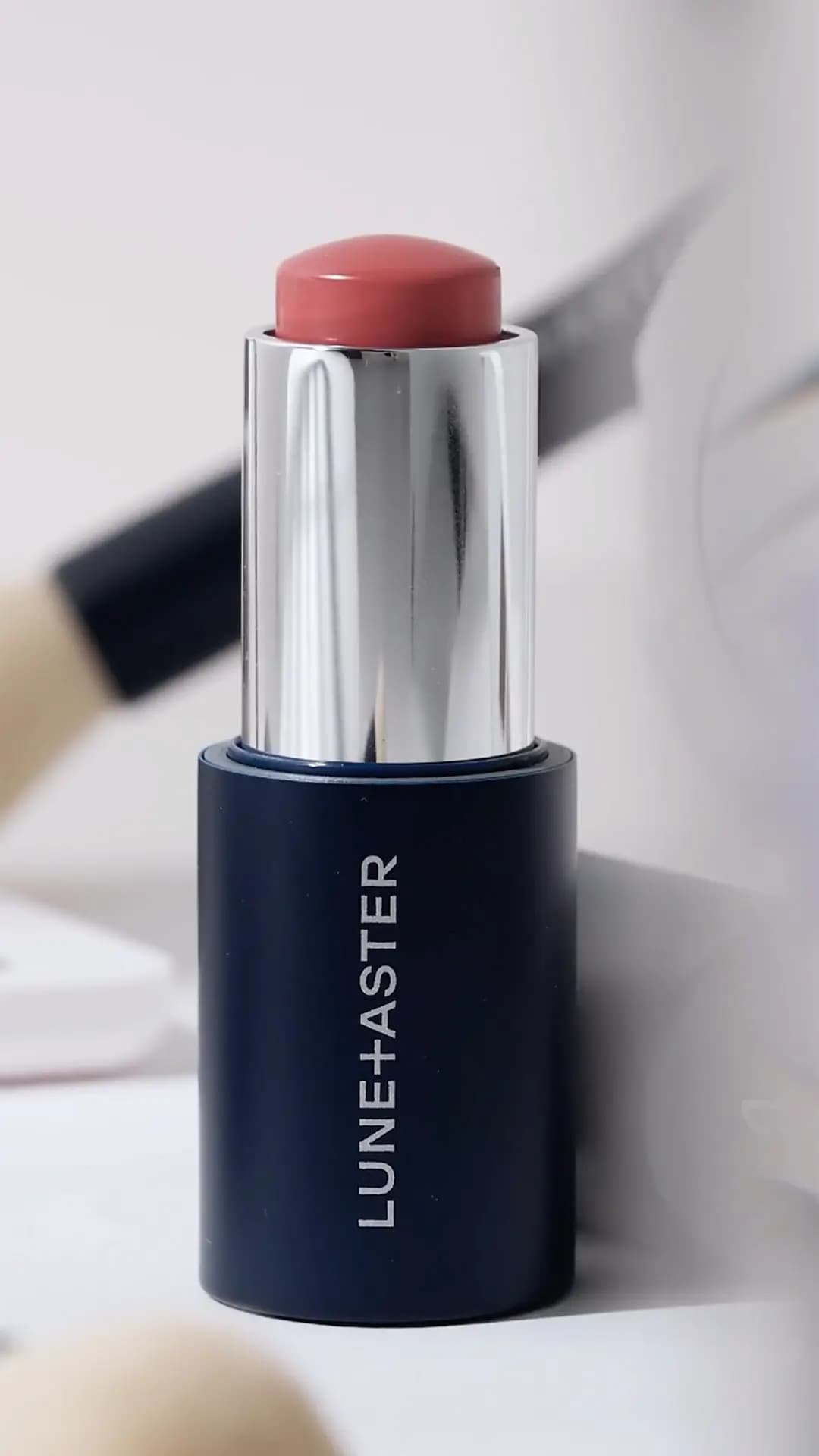 Lune+Aster Cream Multi-Stick in navy-and-silver packaging with a rosy pink bullet, shown against a soft blurred background