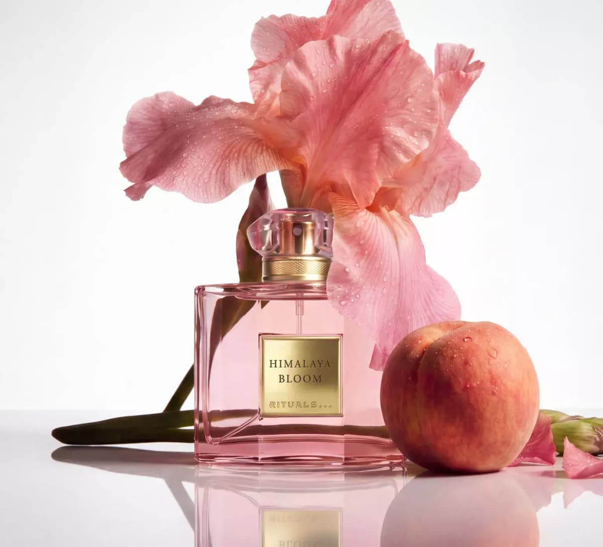Pale pink square Rituals Himalaya Bloom bottle with gold label beneath a dewy pink iris beside a ripe peach