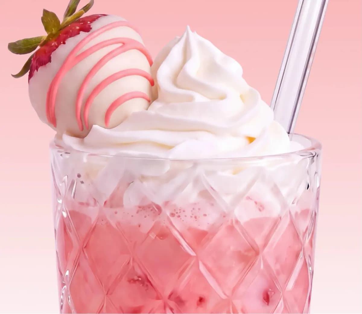 Pink strawberry drink in a diamond-cut glass topped with whipped cream, a white chocolate-dipped strawberry, and a straw
