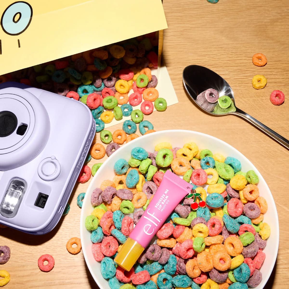 Pink Fruity O's balm resting in a bowl of rainbow cereal beside a lilac instax camera and a spoon