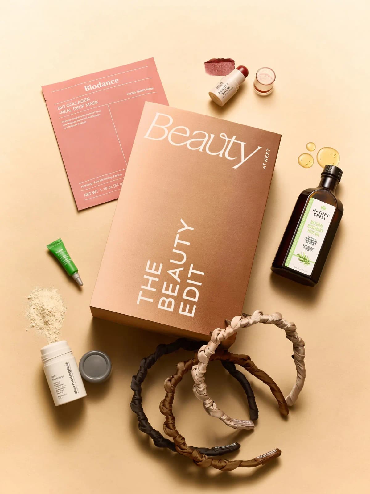 Next The Beauty Edit Box, a bronze gift box surrounded by a sheet mask, balm stick, hair oil, eye tube and headbands